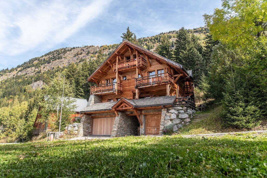 The chalet features a traditional alpine design, showcasing wooden accents and expansive balconies. Surrounded by lush greenery, the structure is positioned against a backdrop of rolling hills and mountainous terrain.