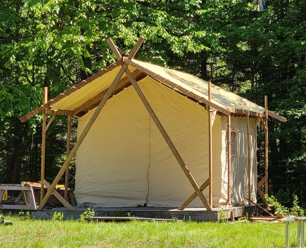 A tent is set within a forest, featuring a sturdy wooden frame and a yellow canvas exterior. The structure is positioned on a wooden deck, surrounded by grass and shaded by trees. Clear visibility of the tent's entrance allows easy access.
