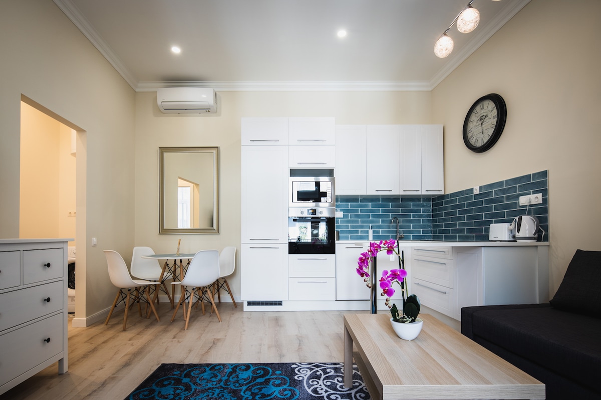 A modern kitchen area is visible, featuring white cabinetry and stainless steel appliances. A dining table with four chairs occupies one corner, complemented by a stylish clock on the wall. A light-colored sofa and a coffee table with a plant create a cozy living space.