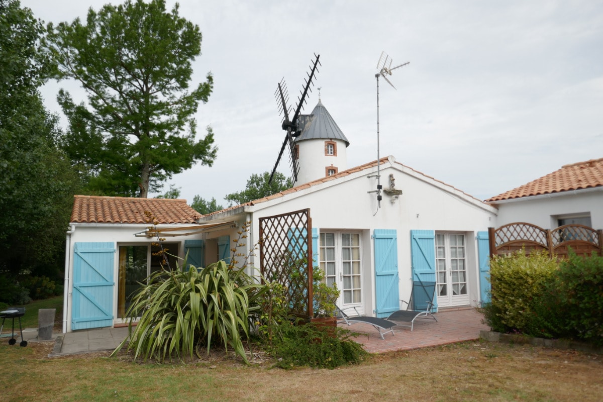 The exterior of a charming cottage features blue-shuttered windows and a terracotta roof. Lush greenery surrounds the building, complemented by a classic windmill in the background, adding character to the landscape. A sunbathing area with loungers is visible on the patio.