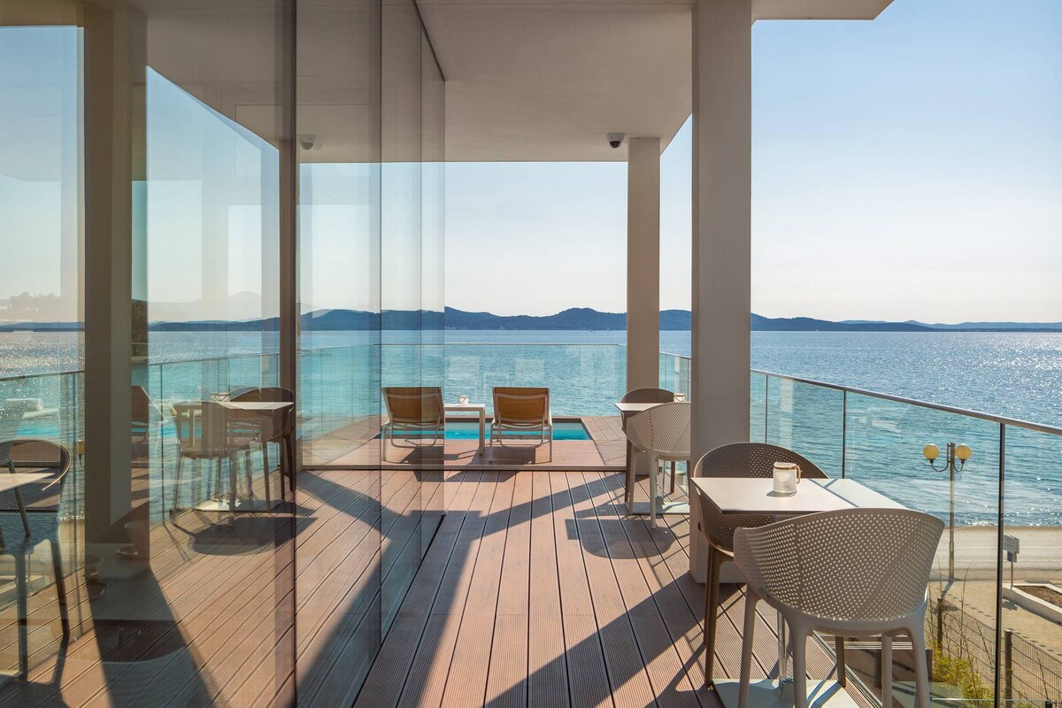 An expansive terrace offers stunning sea views, featuring outdoor seating arrangements with modern chairs and a table. Glass railing enhances visibility of the horizon, while sunlight reflects off the water, creating a serene outdoor space for relaxation.