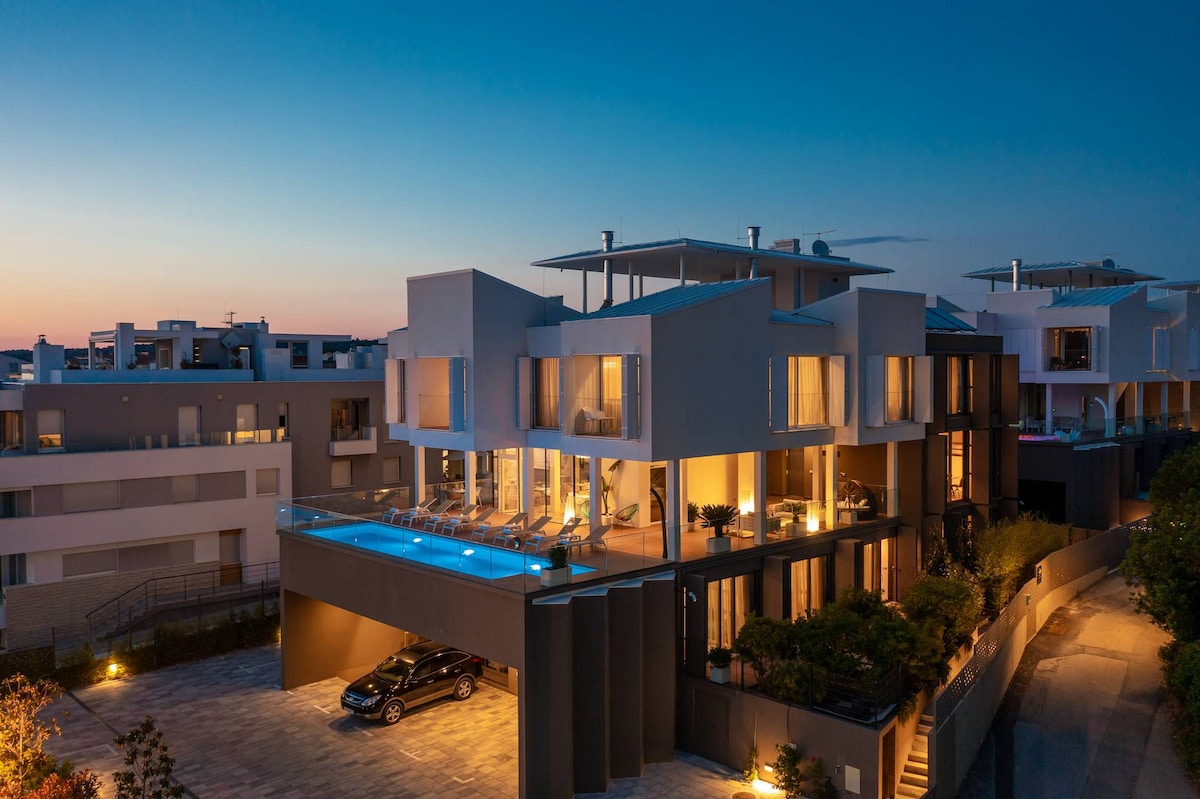 The modern villa is illuminated at dusk, showcasing multiple balconies and expansive windows. A stylish outdoor pool extends along the terrace, with comfortable lounge chairs positioned nearby. The property features landscaped greenery and a parking area, providing a serene and elegant atmosphere.