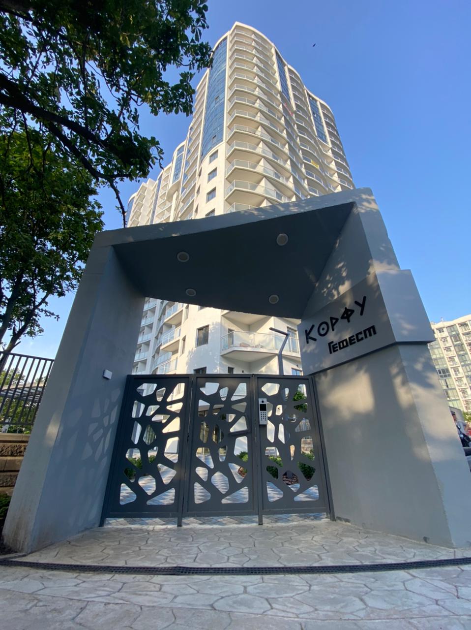 The entrance of a modern residential building is displayed, featuring a contemporary gate with intricate cutout designs. Light shines on the grey stone pathway leading up to the gate, while the tall structure with numerous balconies is visible in the background, framed by green foliage.