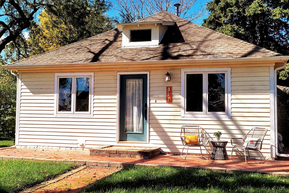 A charming single-story house features a welcoming front porch, framed by large windows that allow natural light to enter. Two outdoor chairs are positioned around a small table on a stone patio, providing a cozy spot to enjoy the surrounding greenery.