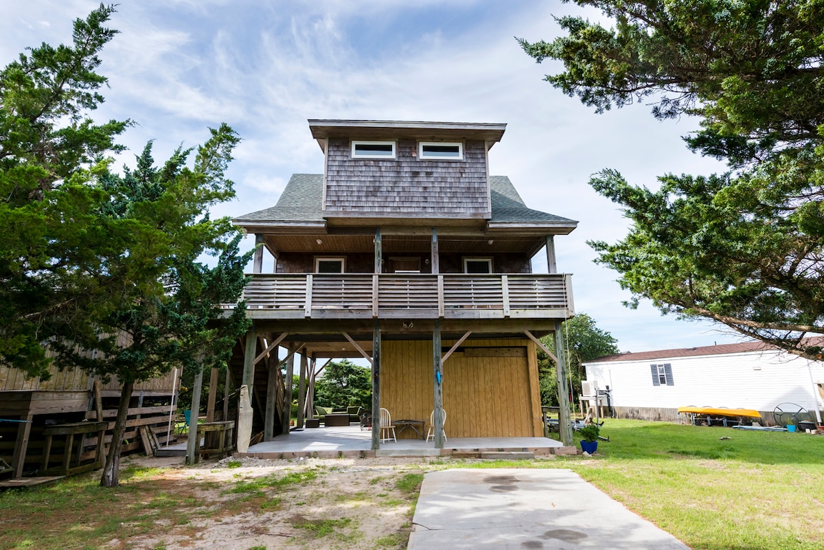A three-story house is set on stilts, surrounded by green grass and trees. A spacious deck with railings extends from the second floor, and the ground level features an open area with a concrete pathway leading to the entrance.