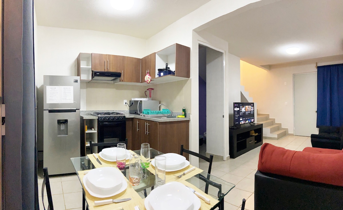 The image shows a modern kitchen adjacent to a living area. A dining table set for four is featured with plates and glasses. Counter space is occupied by kitchen appliances. A sofa and a television are visible in the living area, with stairs leading to the upper floor.
