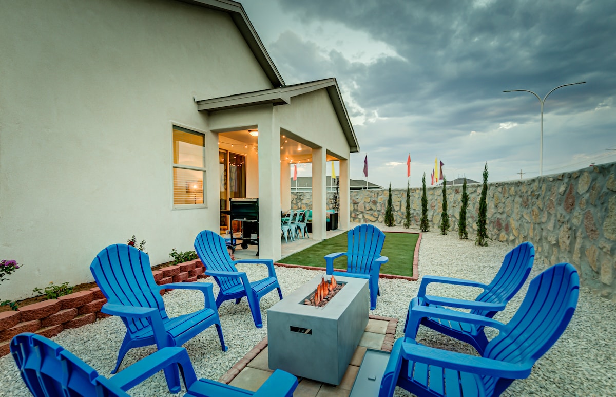 An outdoor space is presented, featuring a fire pit surrounded by six blue adirondack chairs. A well-defined boundary of stones and decorative plants encloses the area, while a covered patio is visible in the background with large windows, contributing to a comfortable gathering environment.