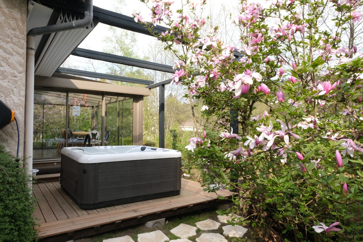 A wooden deck surrounds a well-maintained jacuzzi, positioned under a glass-covered terrace. Lush greenery and blooming pink flowers create a naturally inviting atmosphere. The area blends indoor and outdoor spaces, allowing for a serene retreat amidst the surrounding nature.