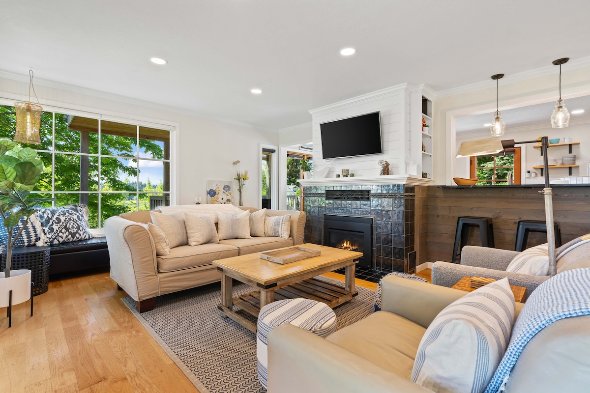 A comfortable living room is showcased, featuring a bright sofa set and a central coffee table. A gas fireplace is positioned beneath a wall-mounted TV, enhancing the cozy atmosphere. Large windows allow natural light to fill the space, offering views of the surrounding greenery.
