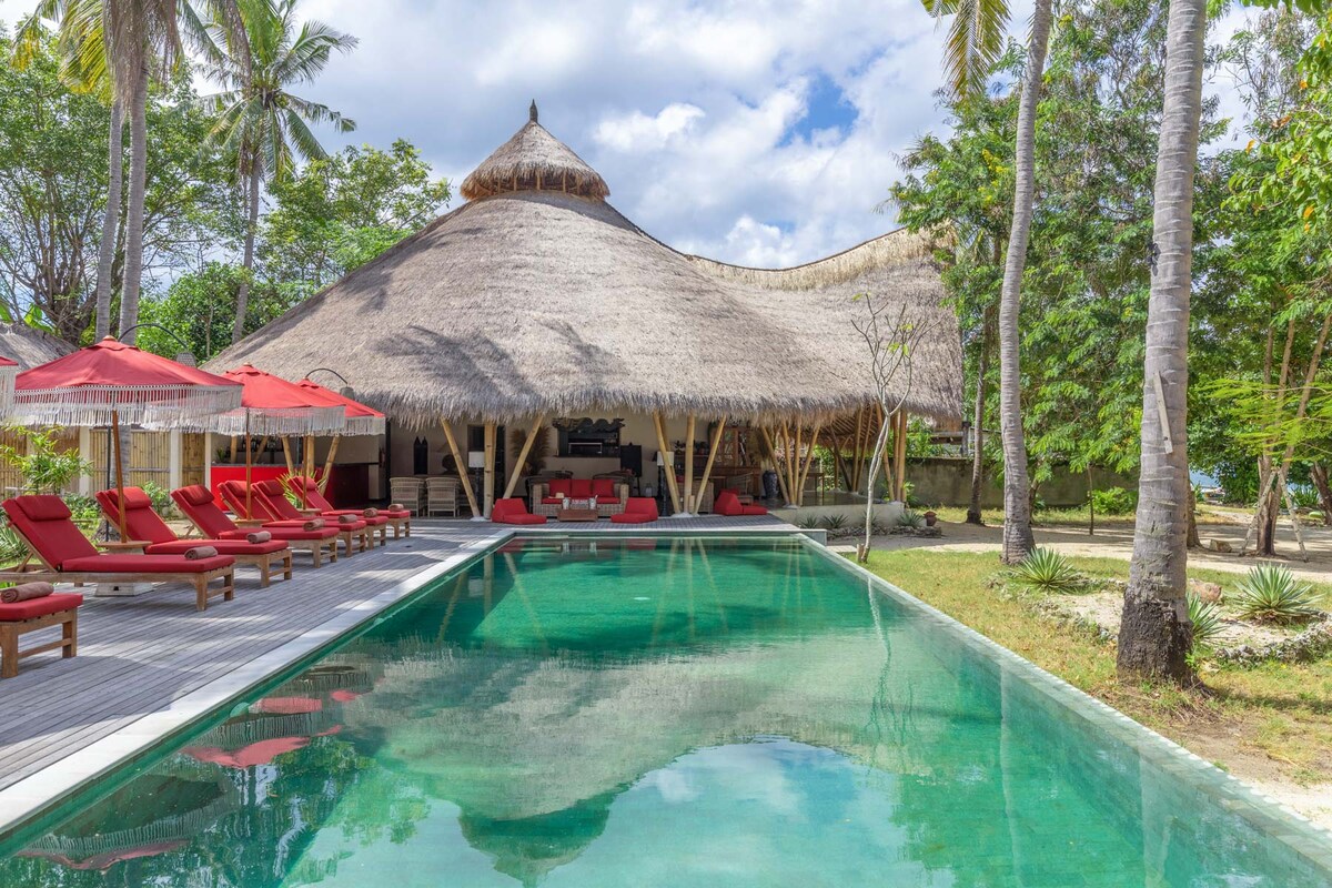A spacious outdoor area is showcased with a serene swimming pool reflecting the sky. Surrounding the pool, red sun loungers provide a comfortable spot for relaxation. The striking alang-alang roof of the villa creates a natural focal point, complemented by palm trees in the background.