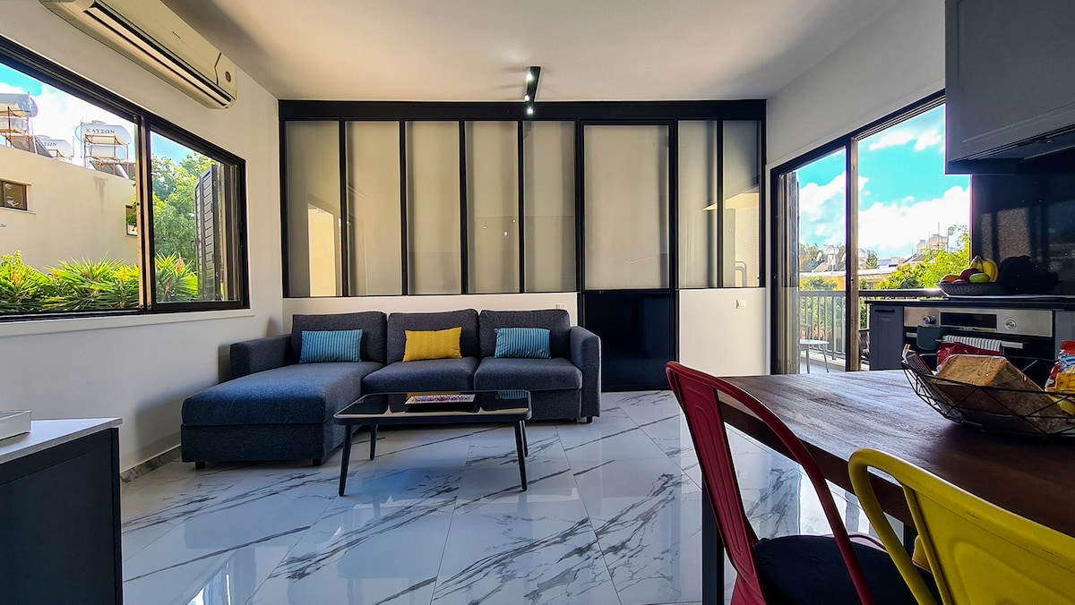 A modern living area is presented, featuring a spacious gray sofa with colorful cushions and large windows that provide natural light. A dining table with red and yellow chairs is visible, along with stylish decor and a well-equipped kitchenette.