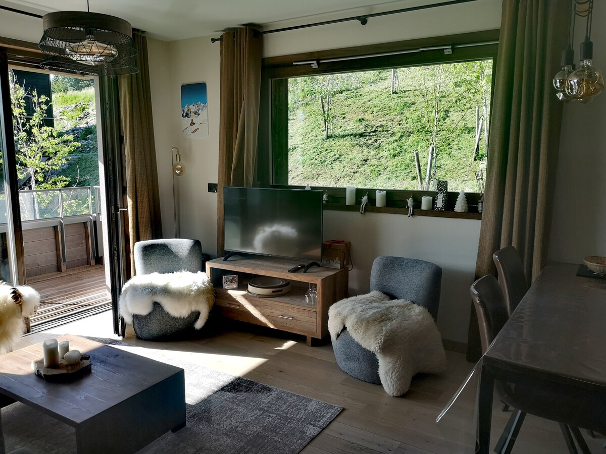 A welcoming living space features a flat-screen television positioned on a wooden unit. Two cozy seating areas with soft throws are present. Panoramic windows provide scenic views of greenery, while natural light fills the room. A dining table is visible in the background.