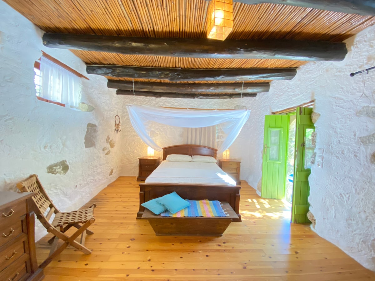 A spacious bedroom showcases a wooden bed with a light coverlet, complemented by soft lighting from two lamps. The natural wood ceiling beams enhance the rustic charm. A vibrant, striped blanket and cushions add color, while a green door provides an inviting entryway.