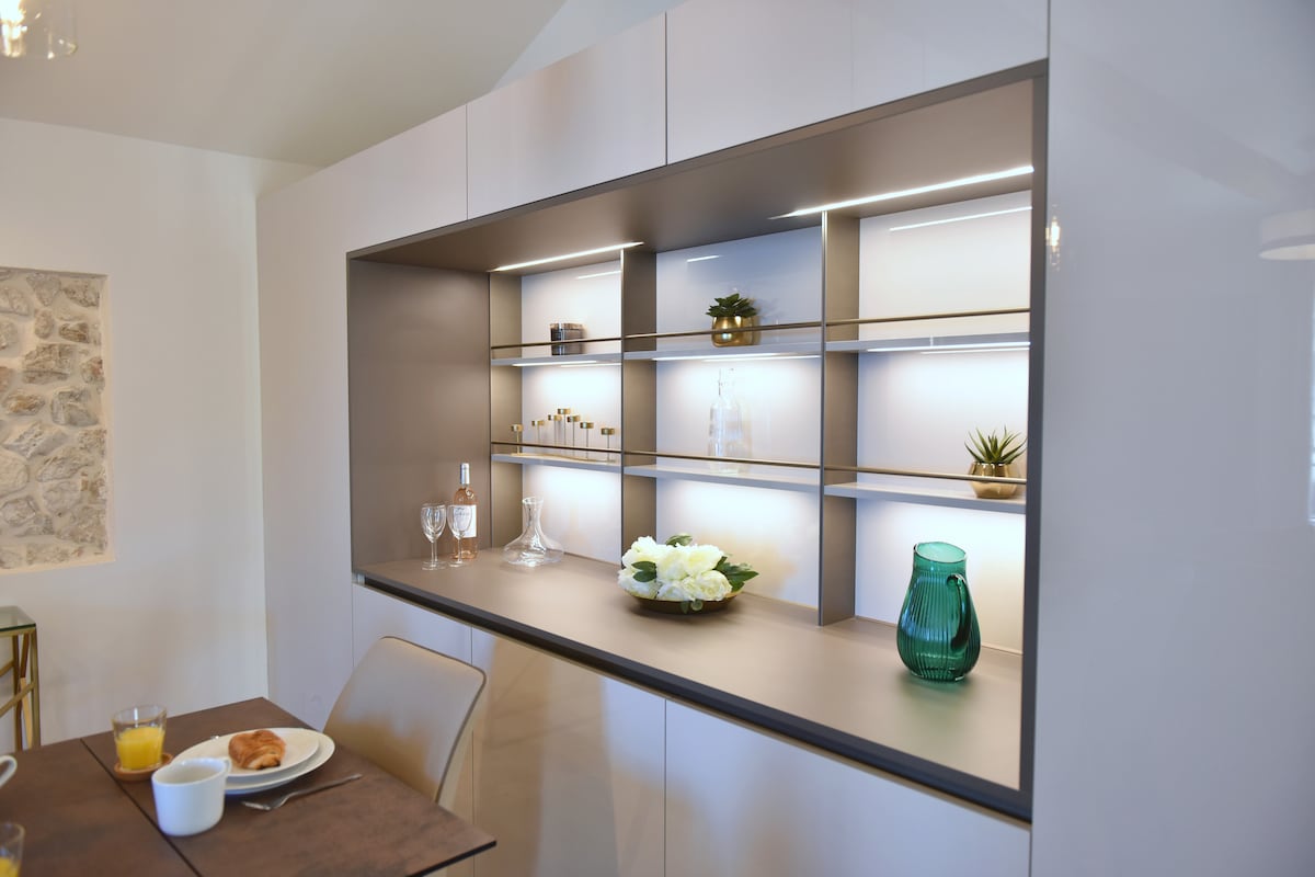 A modern, sleek cabinet with illuminated shelving is showcased. Decorative items, including vases and glassware, are neatly arranged against a backdrop of neutral colors. A dining table is partially visible in the foreground, set with breakfast items, suggesting a welcoming space for meals.
