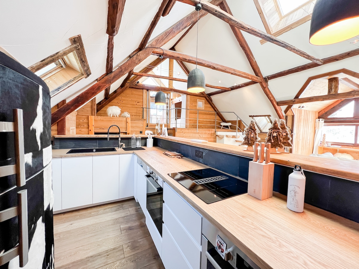 The kitchen features wooden beams overhead, with a spacious countertop and modern appliances. Natural light floods the area through large windows. An inviting island offers additional workspace and seating, complemented by stylish pendant lighting.