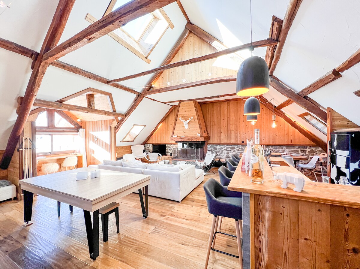 An open living area features a high beamed ceiling, with wooden beams adding a rustic touch. A large dining table is positioned near the center, surrounded by a cozy seating arrangement. Natural light fills the space through multiple windows, enhancing the warm atmosphere.
