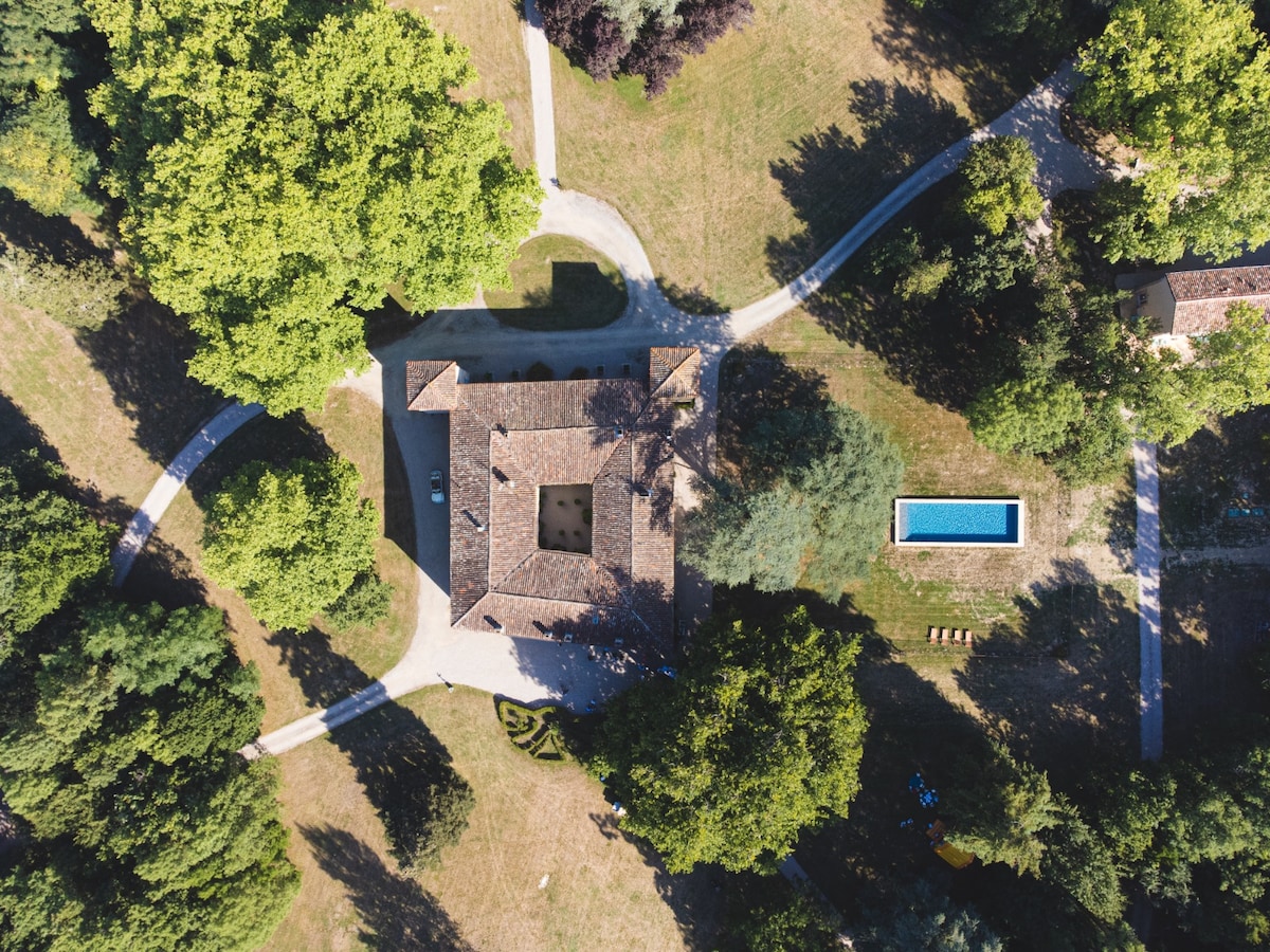 An aerial view displays the property surrounded by lush greenery, with winding paths leading to the main building and a swimming pool. The spacious outdoor area is marked by trees and open grass, creating a serene and natural environment.
