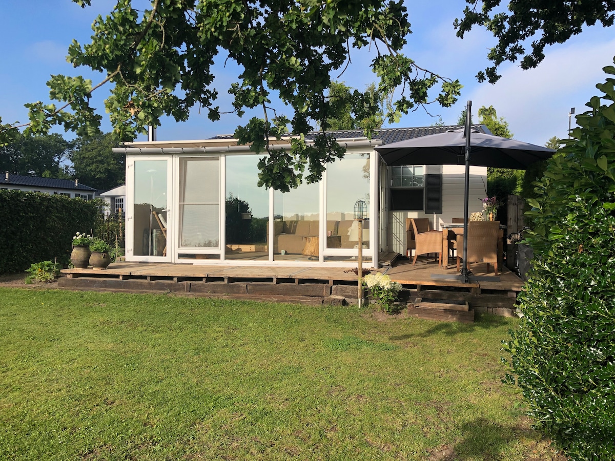The chalet is surrounded by lush greenery, featuring large glass doors that open to a spacious deck. Outdoor seating is provided under an umbrella, inviting relaxation in a tranquil garden setting. This peaceful area is enhanced by natural lighting filtering through the trees.