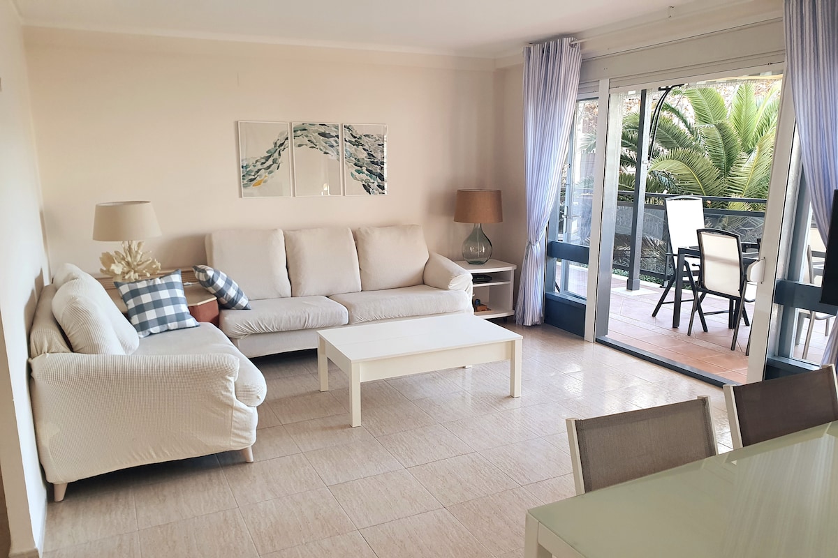 A spacious living area features a light-colored sofa with decorative cushions, a white coffee table, and a floor lamp. Large glass doors provide access to a balcony with outdoor seating, allowing natural light to fill the room. Neutral tones enhance the inviting atmosphere.