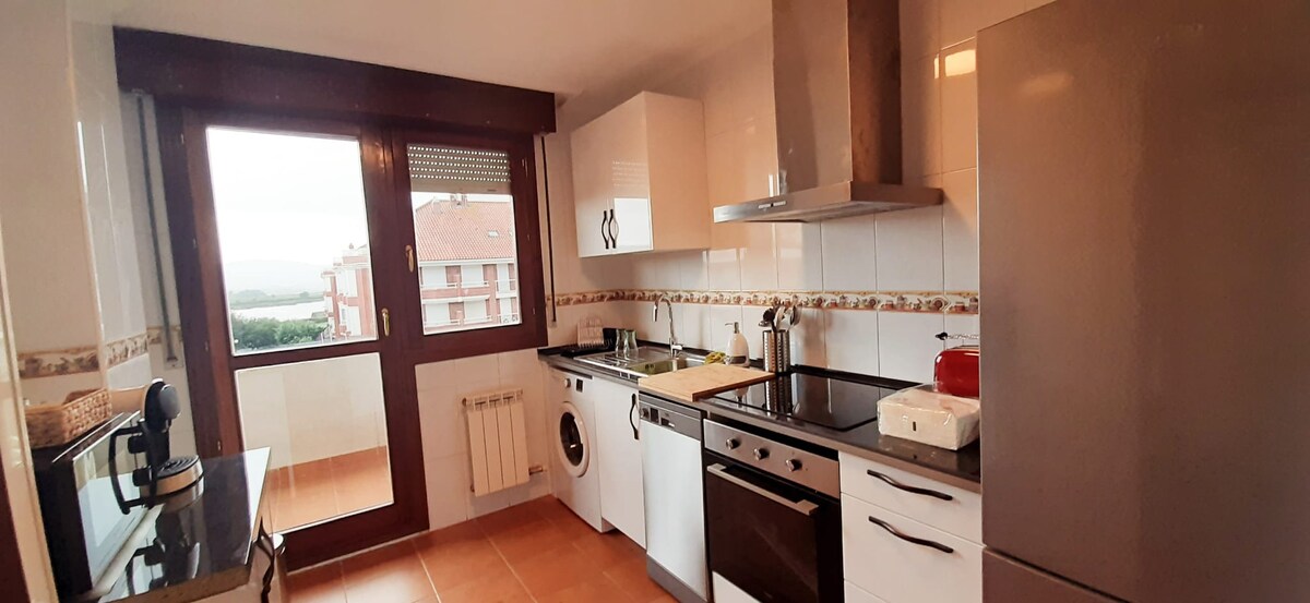 A modern kitchen is showcased with sleek white cabinetry and a combination of stainless steel appliances. A spacious countertop is available for meal preparation, and natural light enters through a large window, offering views of the surrounding area. An adjacent balcony adds extra charm.