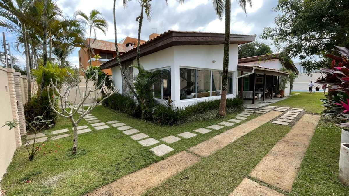 A charming house is surrounded by a well-maintained garden featuring lush greenery and palm trees. A stone pathway leads to the entrance, while large windows offer views of the outdoor space. The exterior exudes a relaxed, beach-inspired aesthetic.