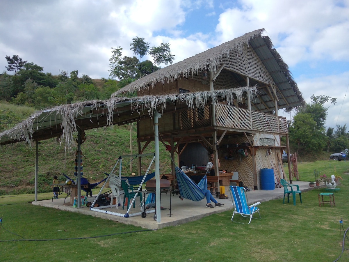 The two-story structure is constructed from natural materials, featuring a thatched roof and open sides. Underneath, various seating options, including colorful chairs and hammocks, are provided. The surrounding green landscape offers a peaceful view, complemented by trees in the background.