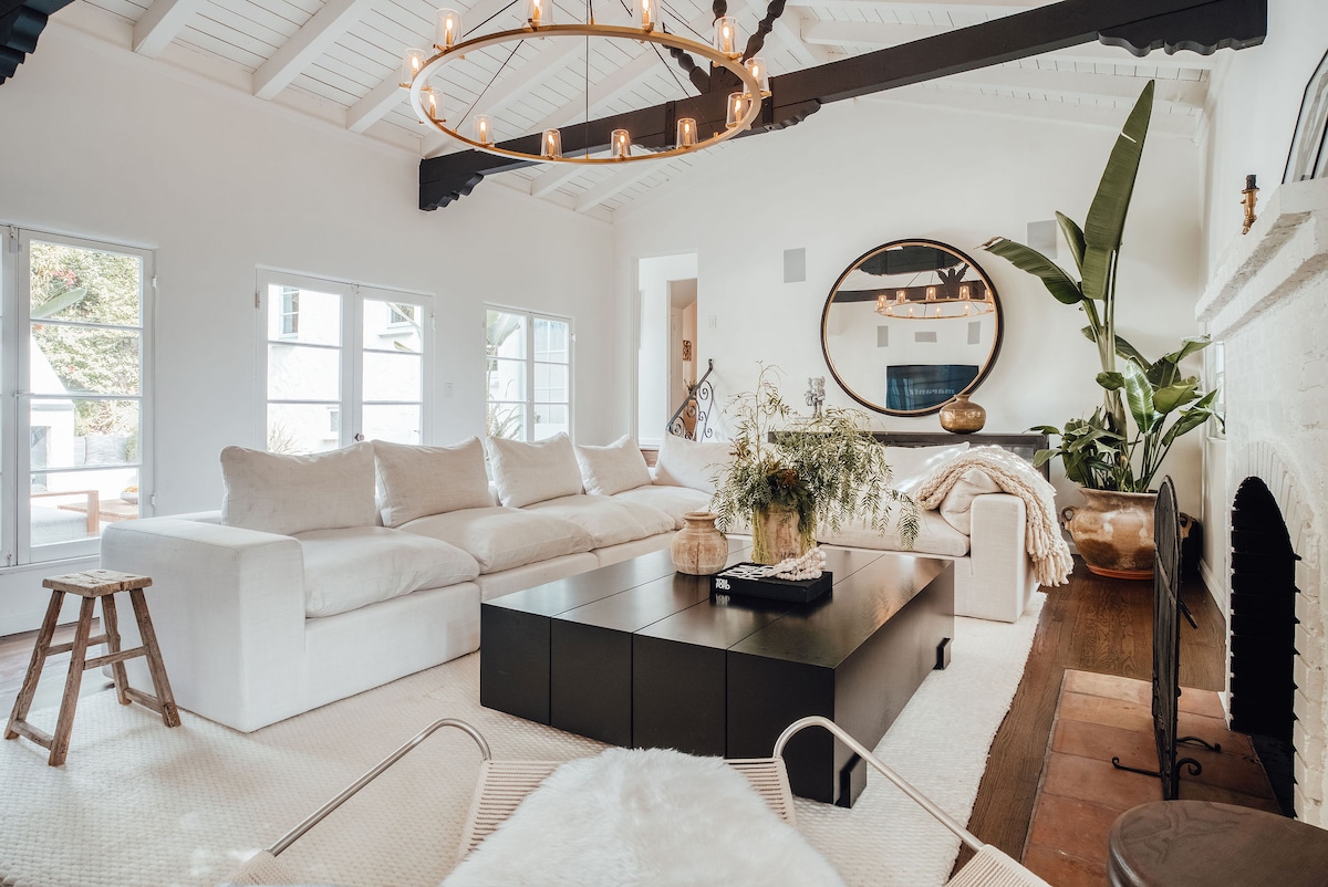 A bright living area is showcased with a spacious sectional sofa and a large coffee table at its center. A large circular mirror reflects natural light, complemented by potted greenery. Multiple windows provide ample brightness, while a cozy fireplace adds warmth to the space.