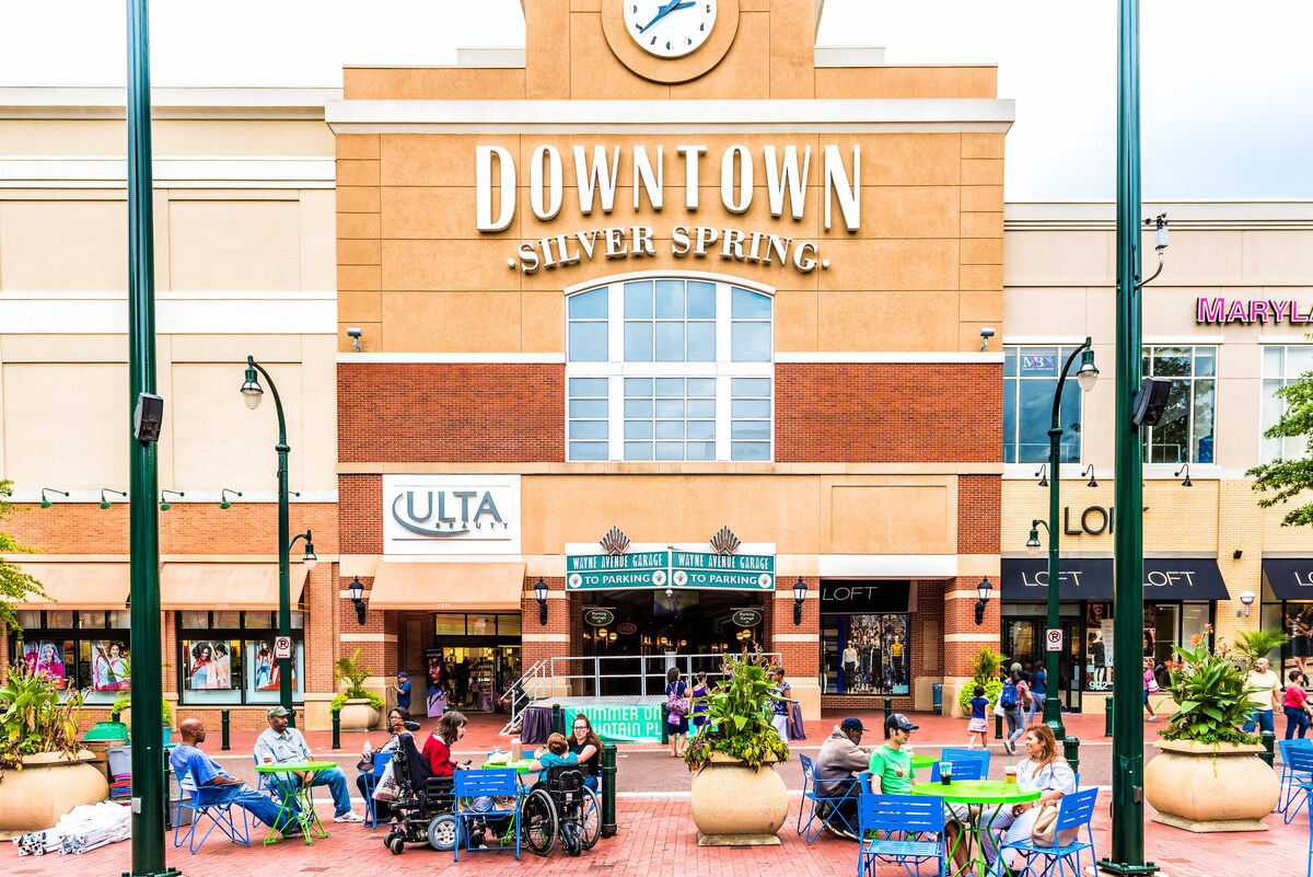 The image depicts a vibrant downtown area in Silver Spring, featuring a large clock mounted on a brick building facade. Pedestrians, including individuals in wheelchairs, are seen enjoying the outdoor seating arrangement with Blue folding chairs. Retail stores, such as Ulta and Loft, are visible.