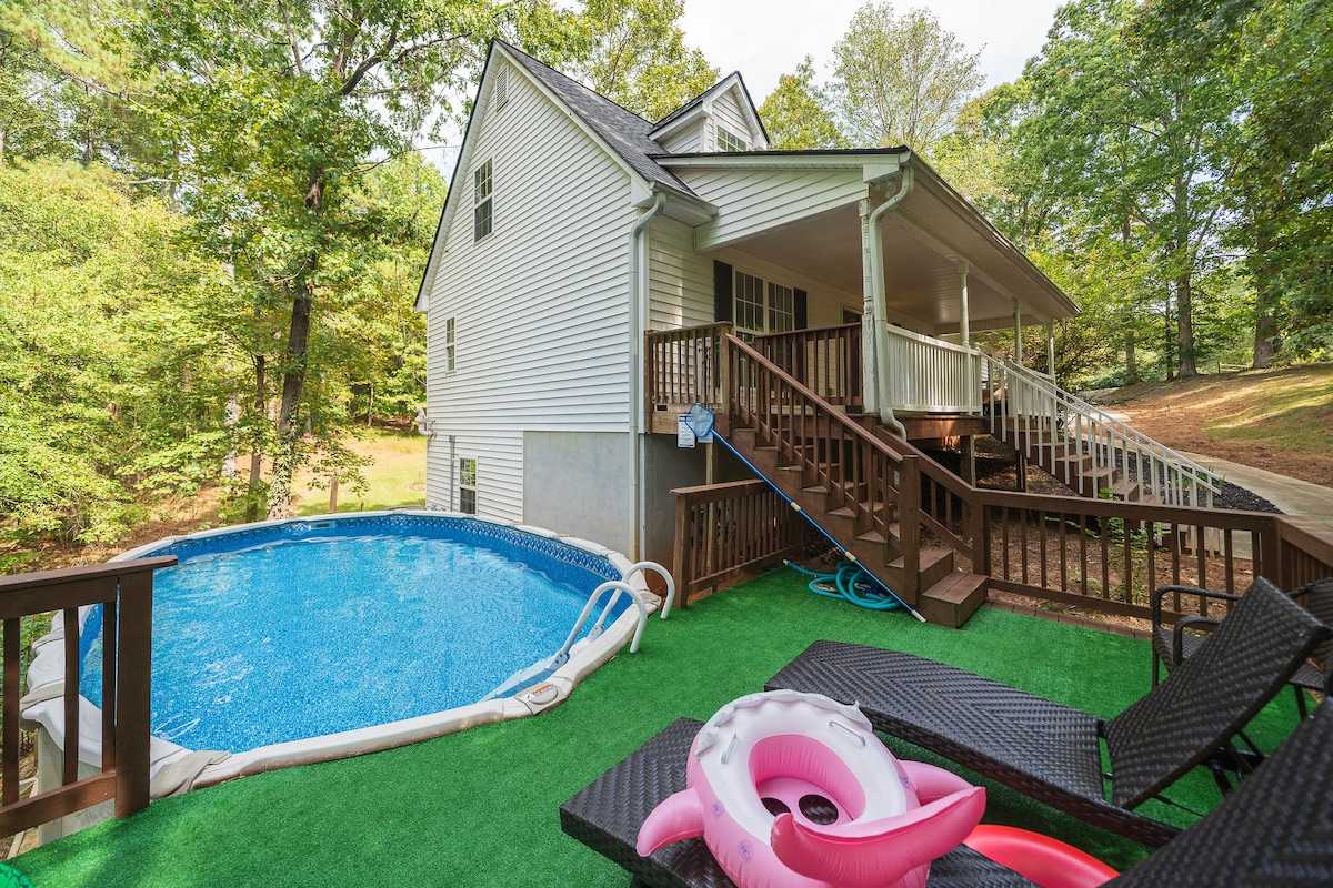 The outdoor area features a private, circular above-ground pool surrounded by a wooden deck. Lounge chairs are positioned nearby, with a pink inflatable float resting beside the pool. The house stands in the background, framed by trees that provide a lush green backdrop.