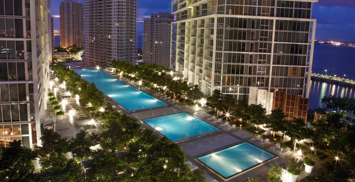 An aerial view captures the illuminated heated pools set amid landscaped greenery, surrounded by high-rise buildings. The soft glow of lights reflects off the water, creating a serene atmosphere. The backdrop includes a glimpse of the waterfront and nearby city skyline at twilight.