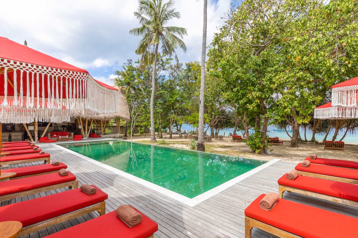 A tranquil view of a pool surrounded by vibrant red loungers, situated within a lush garden. Palm trees and chairs are visible, offering a serene space for relaxation. The clear water reflects the sky, enhancing the inviting atmosphere of the outdoor setting.