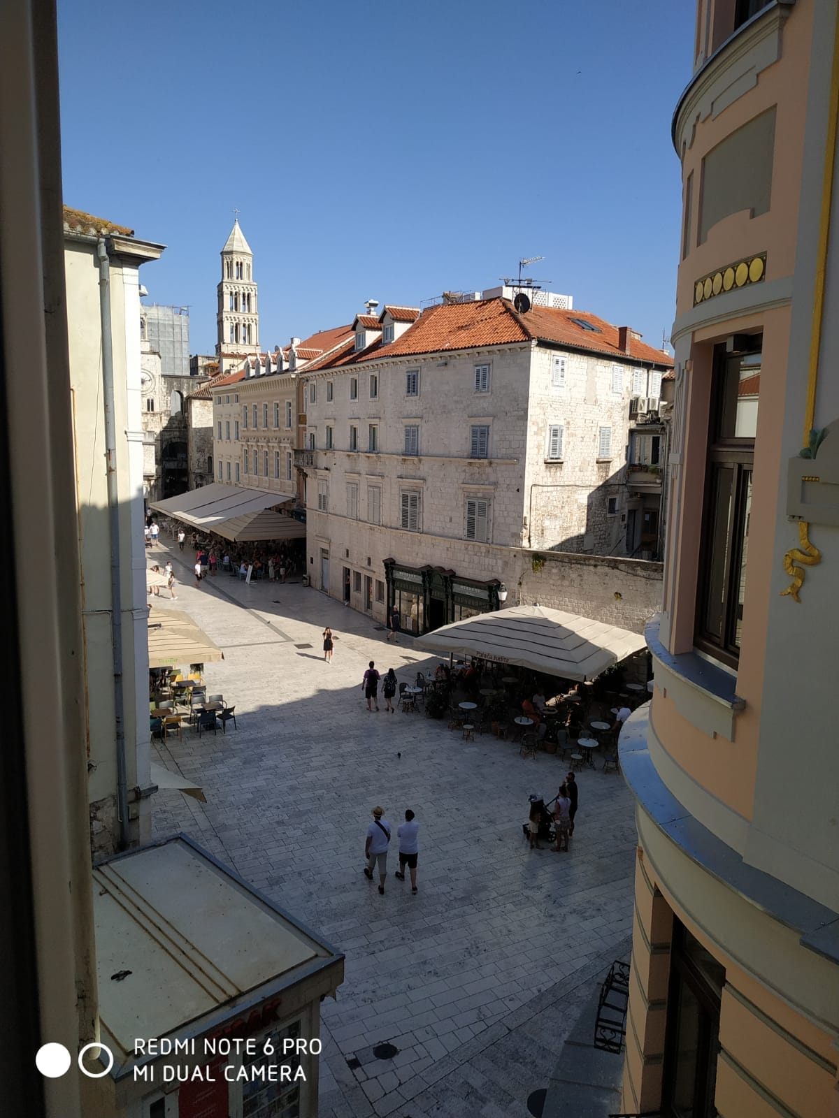 A view of the bustling square, featuring outdoor seating arrangements beneath awnings. Historic buildings frame the scene, with a tall bell tower visible in the background. People are seen strolling along the cobblestone streets, creating a lively atmosphere.