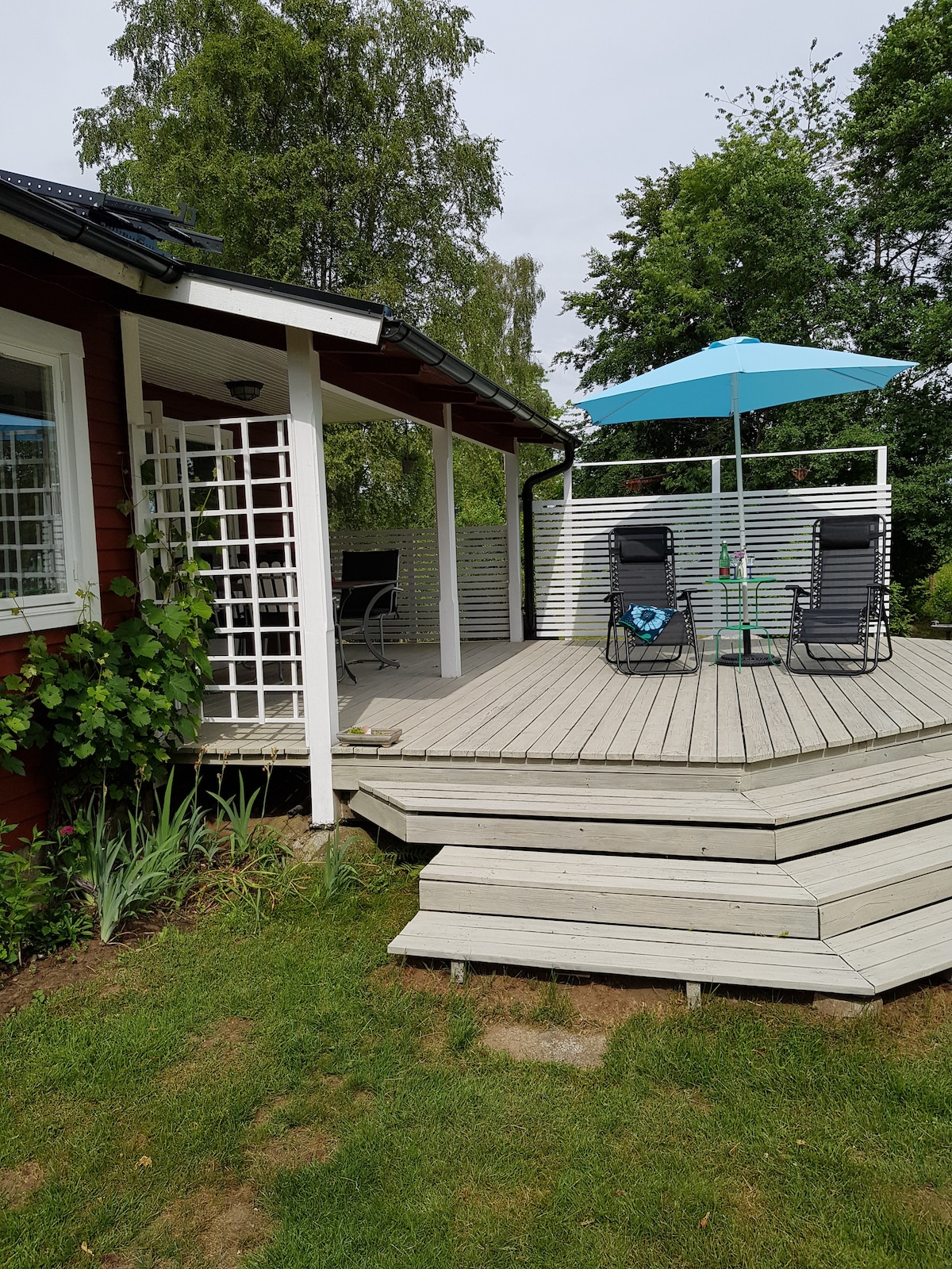A spacious wooden deck extends from the cabin, featuring a patio umbrella over a dining table and several chairs. Lush greenery surrounds the area, enhancing the natural setting. A lattice structure on the side offers additional charm to the outdoor space.