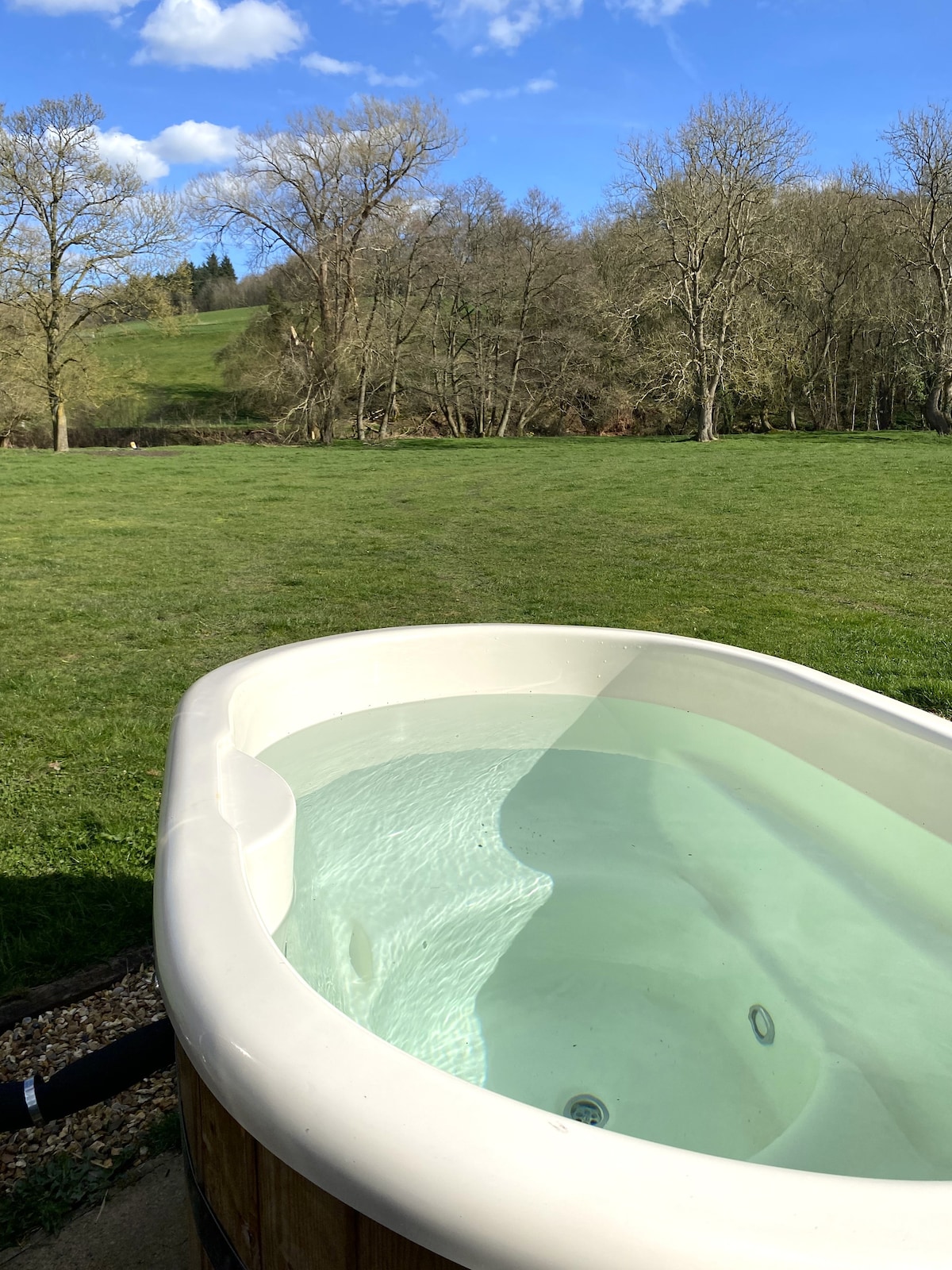 A wood-fired hot tub is partially visible, featuring clear water reflecting the blue sky. In the background, a grassy field leads to wooded areas and rolling hills, creating a serene outdoor setting.