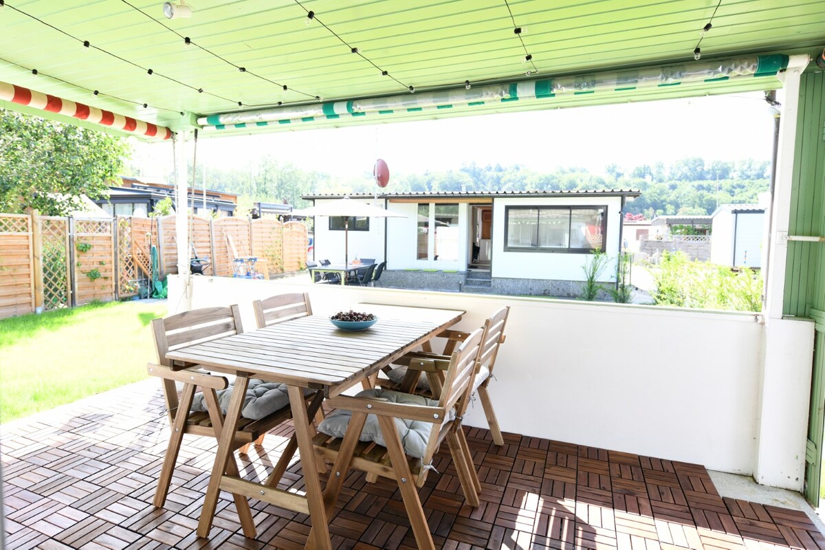 A covered terrace features a wooden dining table surrounded by several chairs, offering a view of the garden and neighboring accommodations. Sunlight filters through the green roof, and the tranquil space invites outdoor dining and relaxation.