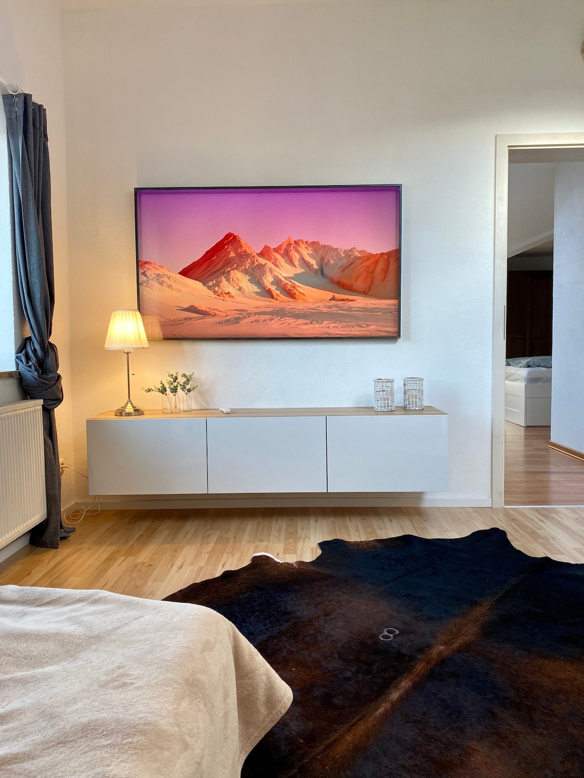 A modern living space is showcased with a sleek white sideboard beneath a large framed artwork depicting a mountain landscape in warm tones. A cozy lamp provides soft lighting, while a decorative arrangement of flowers adds a touch of elegance. A soft, dark rug complements the wooden floor.
