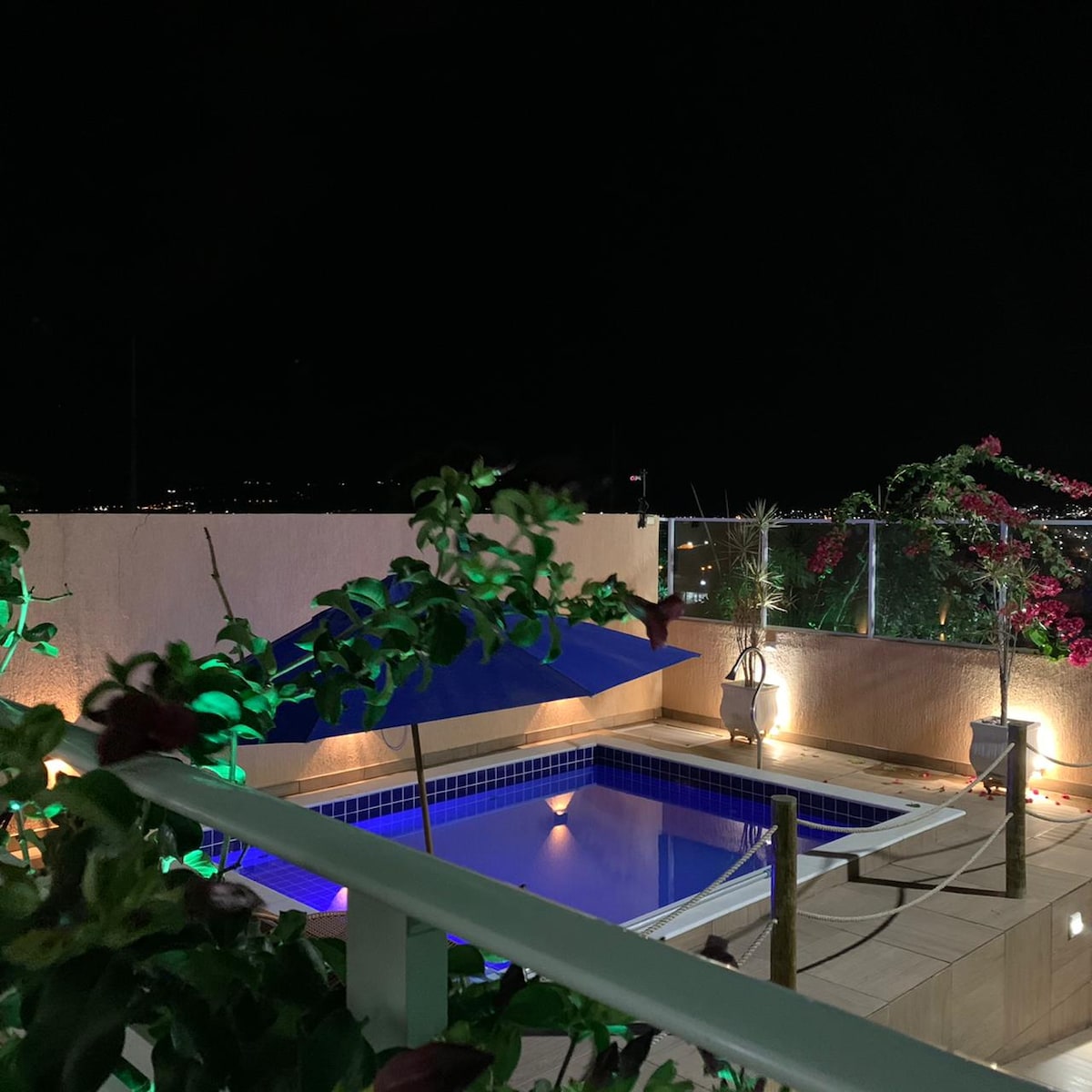 A private pool is framed by lush greenery and soft lighting, creating a serene ambiance at night. The pool area features a shaded seating space, while decorative plants add natural beauty. The view of distant lights enhances the tranquil setting.