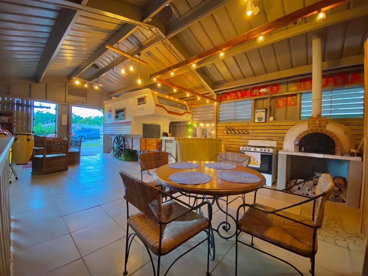 An open kitchen area features rustic wooden details and a circular dining table surrounded by four wicker chairs. Soft ambient lighting highlights the space, with a traditional oven and grill set nearby. Large windows provide a view of the surrounding nature.