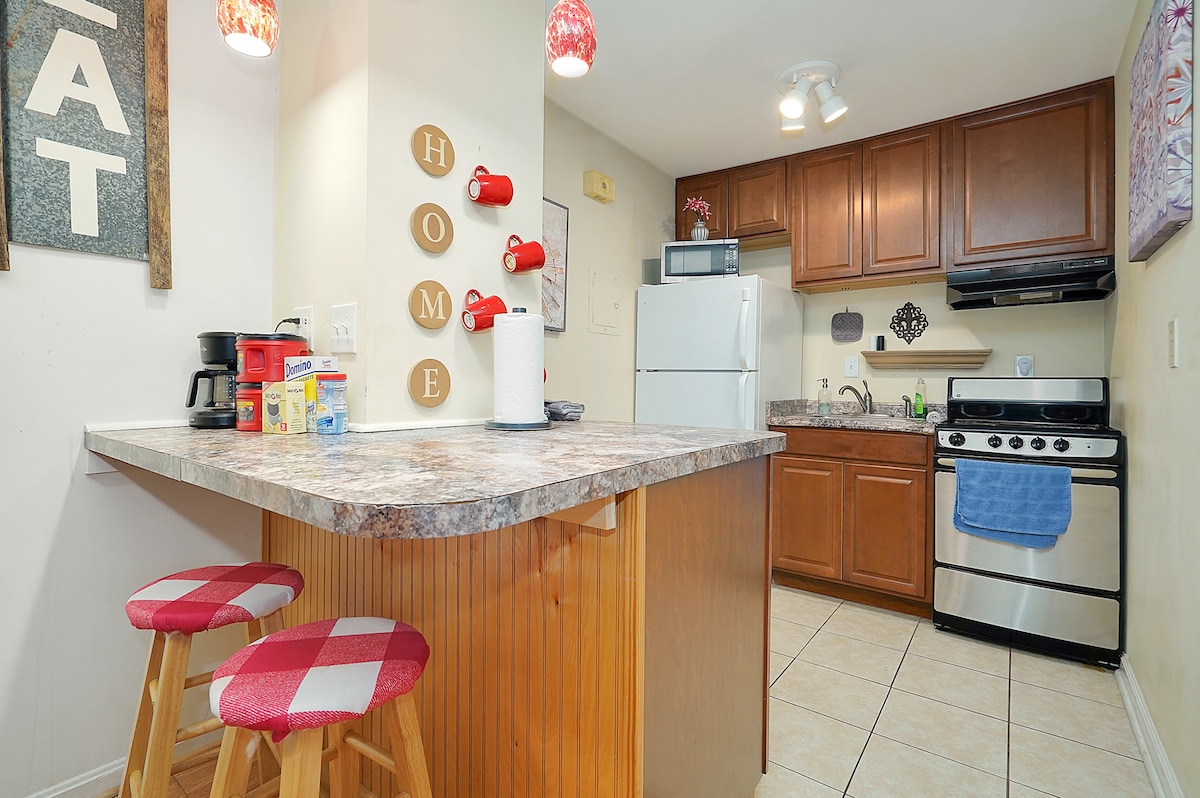 A compact kitchen area is showcased with warm wooden cabinetry and light-colored tile flooring. A round bar counter with two red and white checkered stools is visible, along with essential appliances, including a refrigerator and stove. Decorative elements include wall-mounted red mugs and 'HOME' letters.