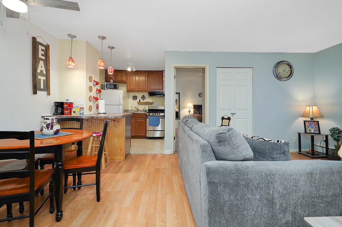 A welcoming living space is presented, featuring a comfortable gray sofa facing a wall-mounted clock and a decorative piece. A cozy dining area with a circular table contrasts with the light wood floor. Bright pendant lights above enhance the kitchen's warm ambiance.