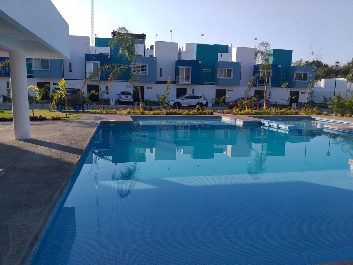 A clear swimming pool reflects the surrounding buildings and lush landscaping. Colorful structures are visible in the background, complementing the serene outdoor space. Palm trees and well-maintained gardens enhance the inviting atmosphere. The scene is calm, emphasizing a relaxing environment for guests.
