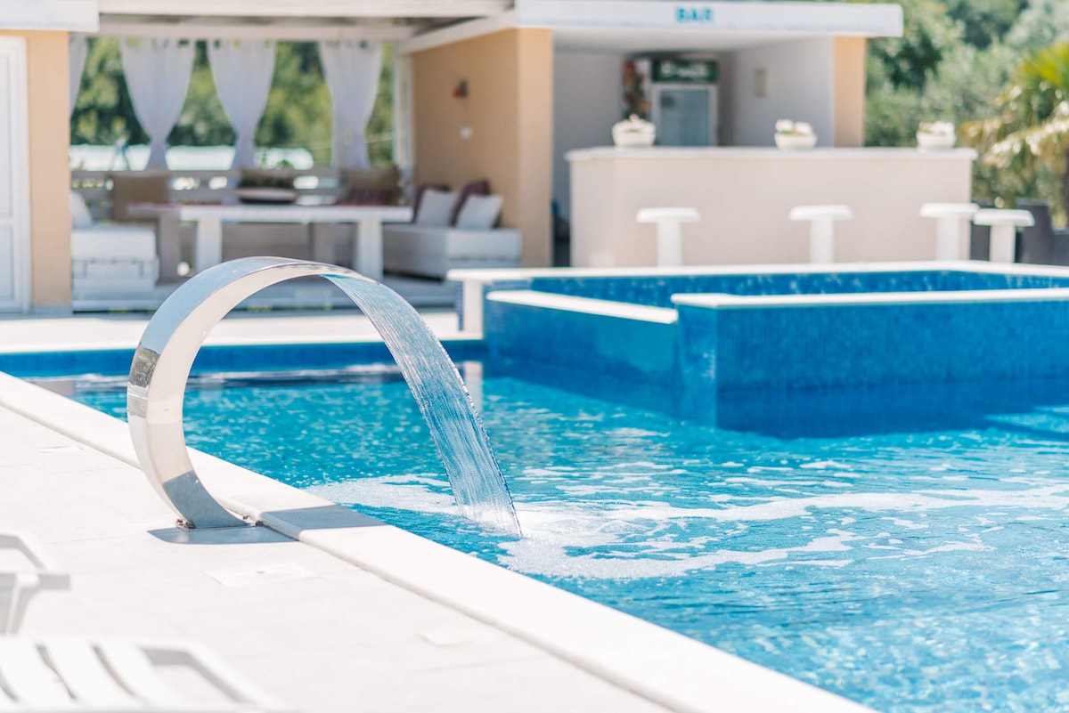 A modern swimming pool is featured, with a distinctive curved water feature creating a gentle flow into the clear blue water. Sun loungers are positioned nearby, and a bar area is visible in the background, surrounded by greenery.