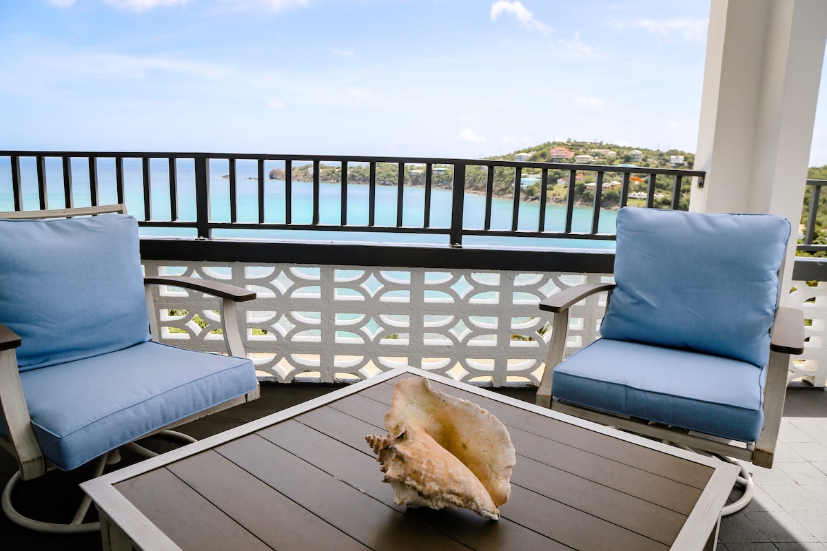 A serene outdoor seating area is presented on a balcony, featuring two light blue chairs beside a dark wooden table. An attractive seashell rests on the table, while a panoramic view of the calm, blue waters and distant hills is framed by a decorative railing.