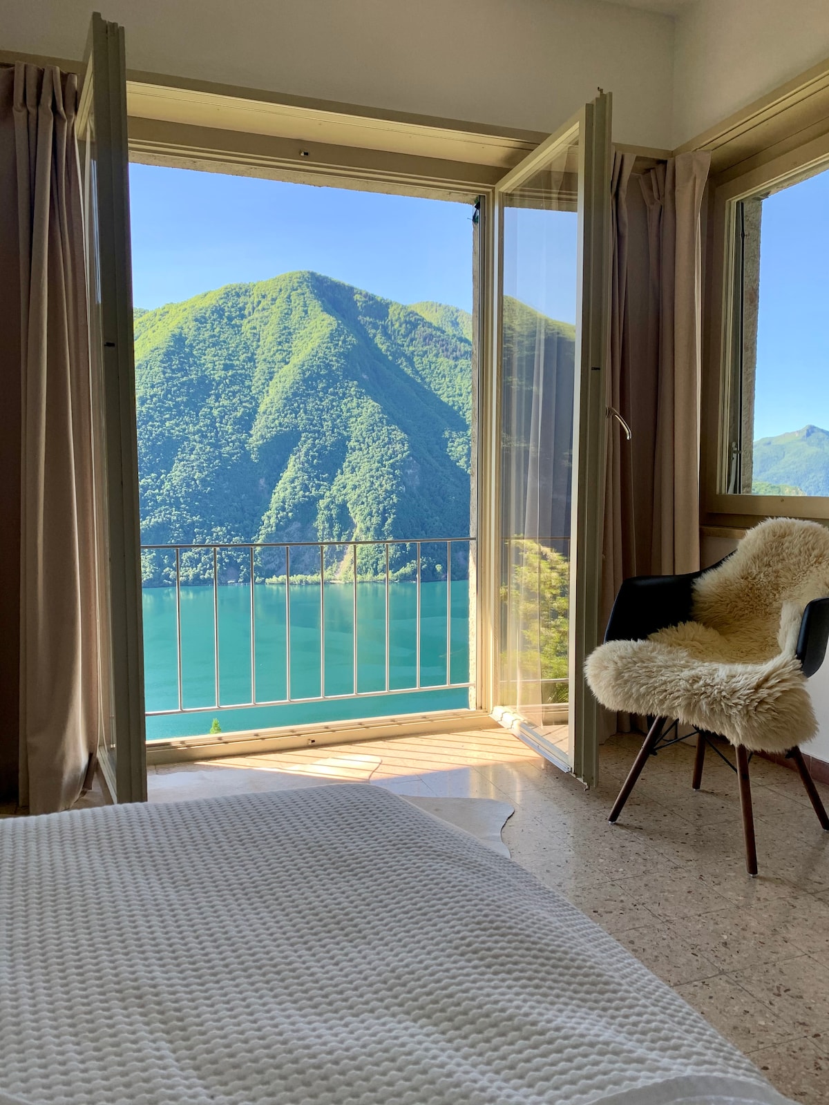 A tranquil bedroom features large open doors leading to a stunning mountain view. Soft light fills the space, highlighting the textures of a cozy quilt on the spacious bed. A fur-covered chair adds a touch of comfort, enhancing the serene ambiance.
