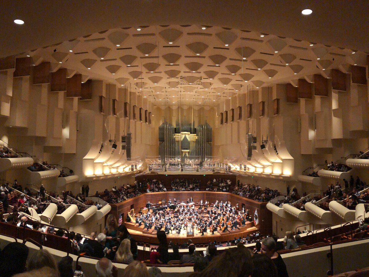 An interior view of a concert hall showcases a modern architectural design, featuring a towering organ at the back. Seating is arranged in a circular formation around the stage, which is filled with musicians. The ceiling is adorned with an intricate pattern, creating an engaging visual effect.