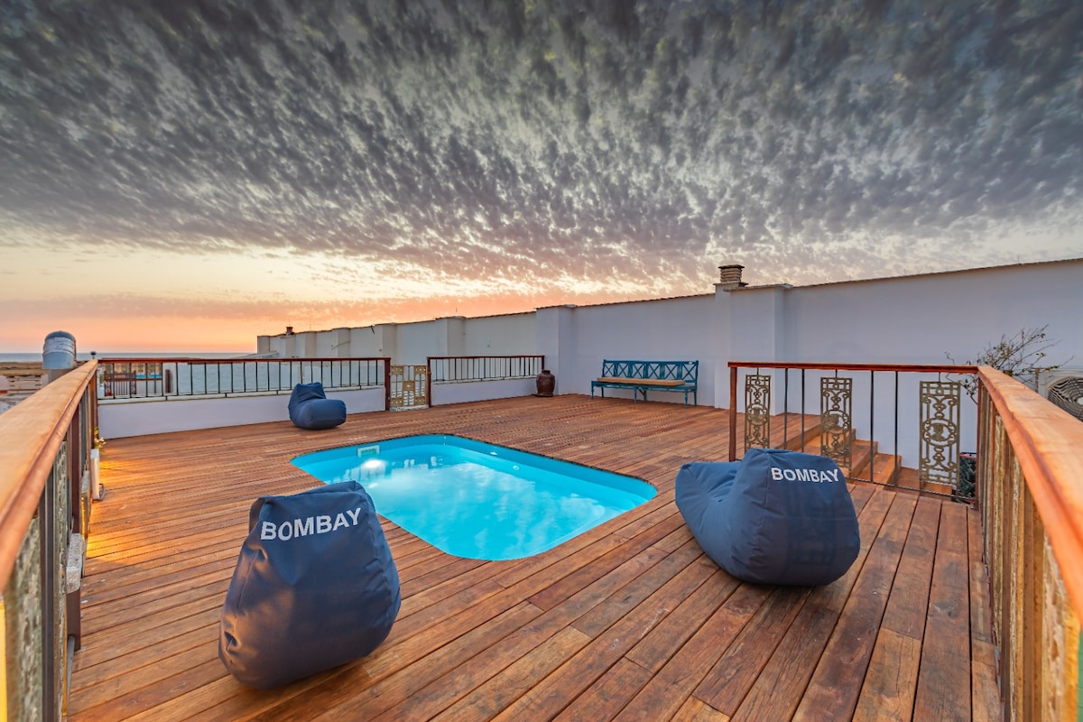 An outdoor terrace features a wooden deck surrounding a small swimming pool. Two comfortable bean bag chairs are positioned beside the pool, offering a place to relax. The sky displays a mixture of soft colors as the sun sets in the background.
