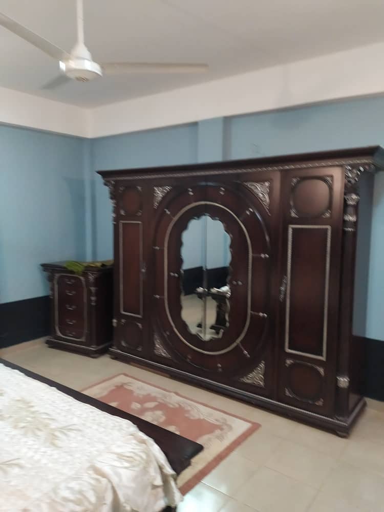 A room features a large, ornate wooden wardrobe and a matching nightstand. The walls are painted in a soothing blue, and a simple area rug lies on the tiled floor. A ceiling fan is positioned above, providing air circulation.