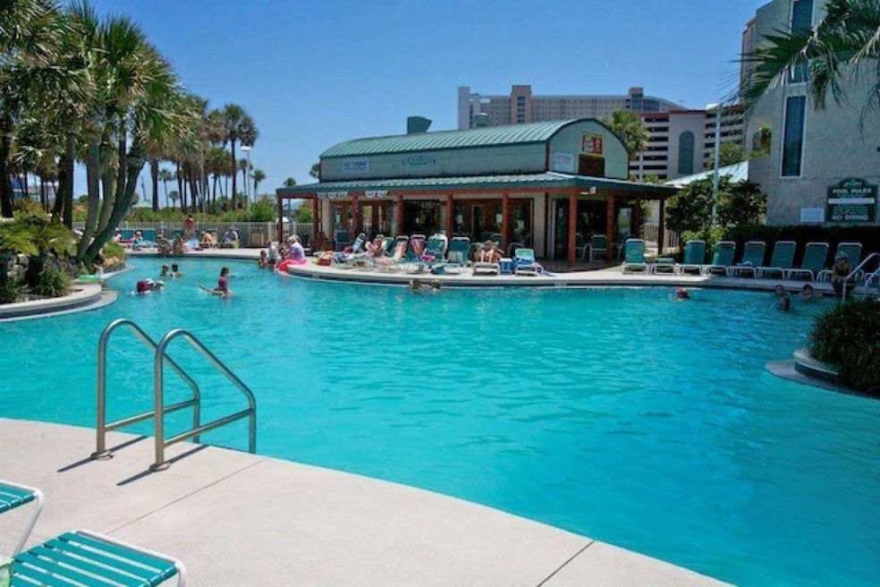 A lagoon-style pool features clear turquoise water and palm trees, creating a tropical atmosphere. A poolside bar is visible, with guests enjoying refreshments. Lounge chairs are arranged around the pool, providing ample seating for relaxation. Sunshine casts a bright glow, enhancing the inviting scene.