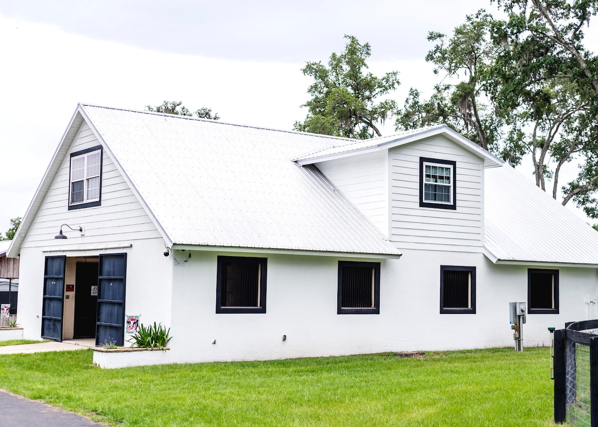 A charming barn structure, featuring a white exterior and a distinctive gabled roof. Two large blue double doors create an inviting entryway, flanked by several windows that allow natural light to fill the interior. Lush green grass surrounds the building, contributing to the serene environment.