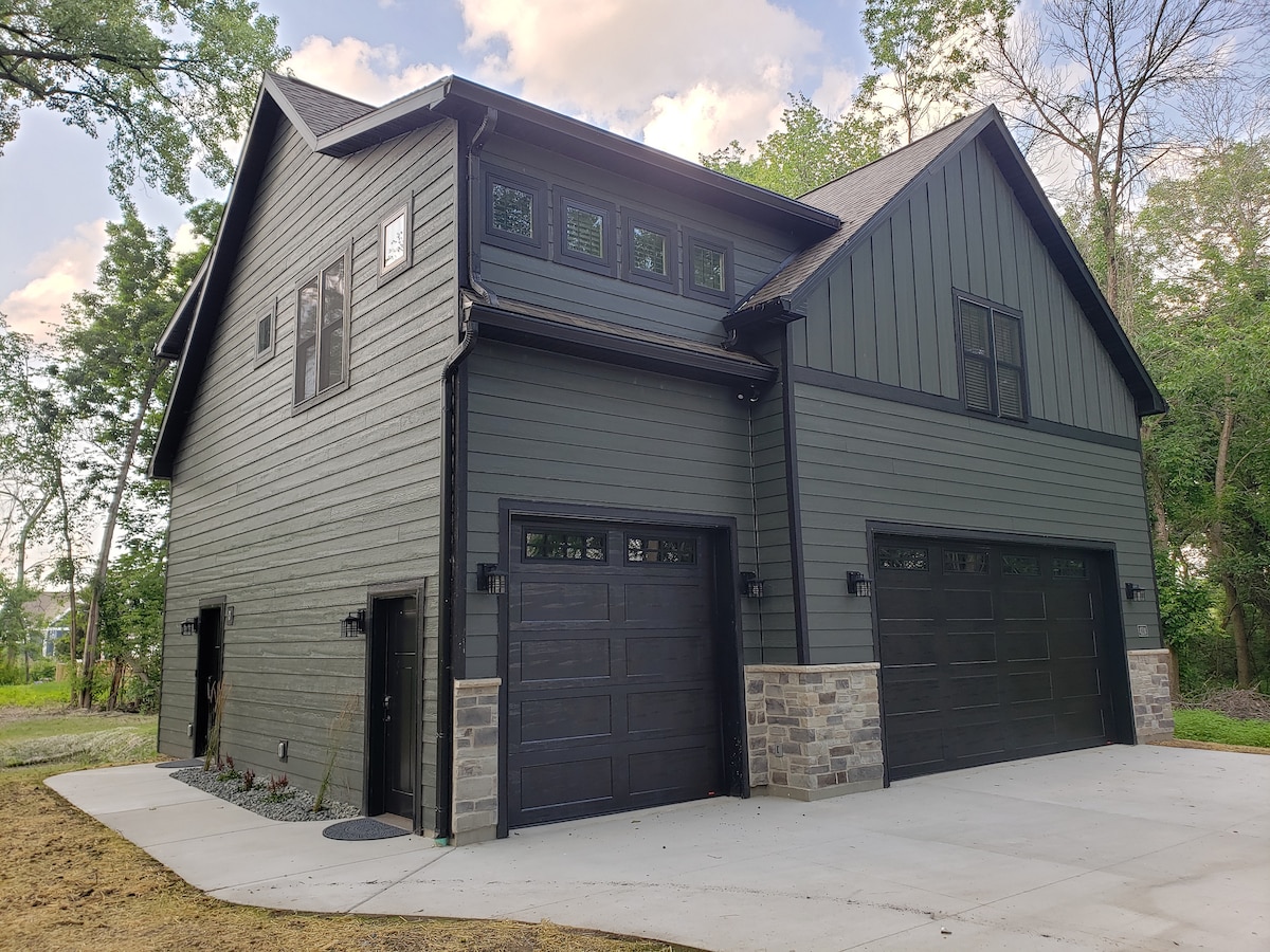 The exterior of the home showcases a modern design with dark green siding and stone accents. Two garage doors are visible, complemented by decorative light fixtures. A concrete driveway leads to the entrance, surrounded by landscaped areas and greenery.
